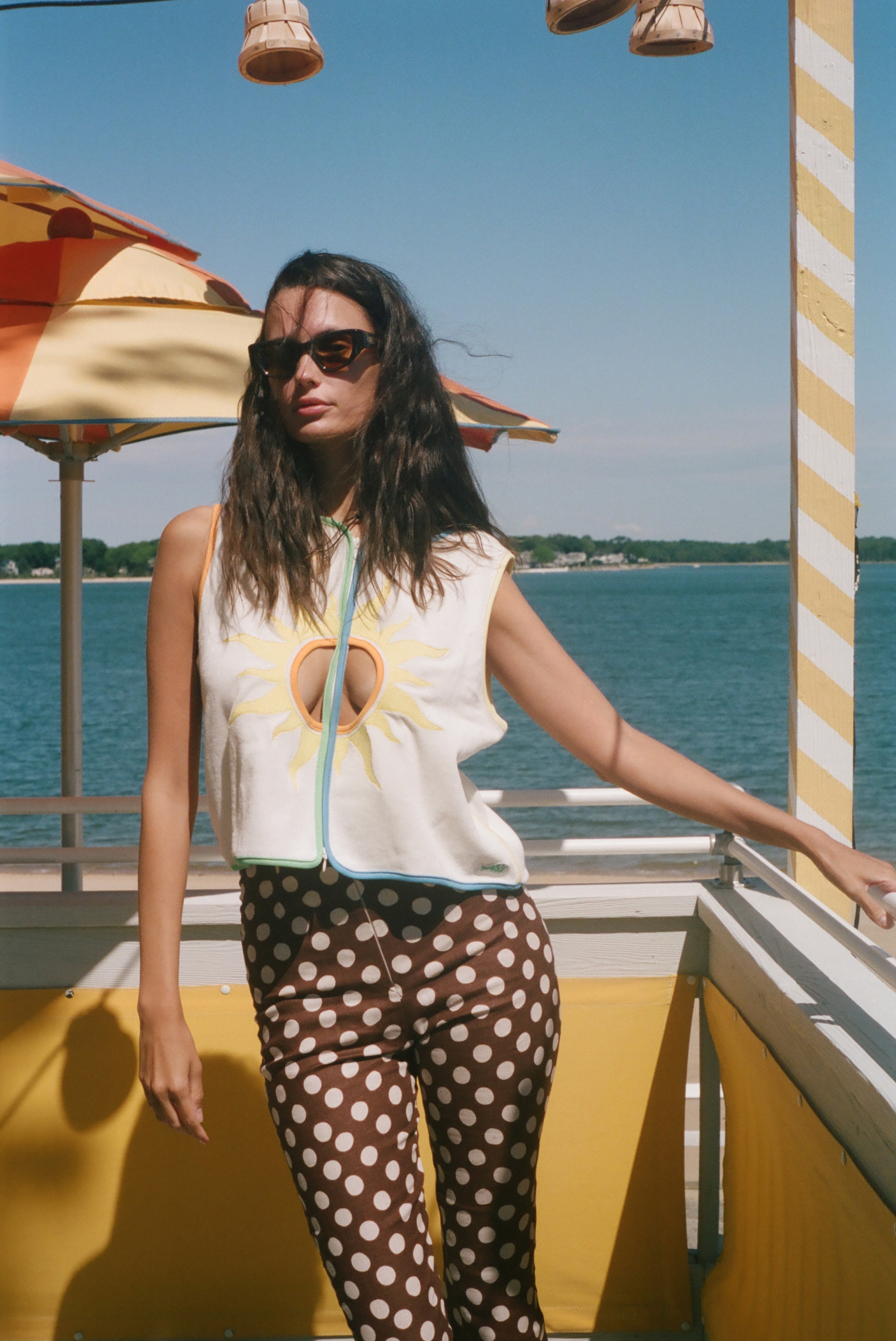A woman wearing a sleeveless white top with a yellow sun graphic and brown polka-dot pants stands on a sunny outdoor deck with water and umbrellas in the background.