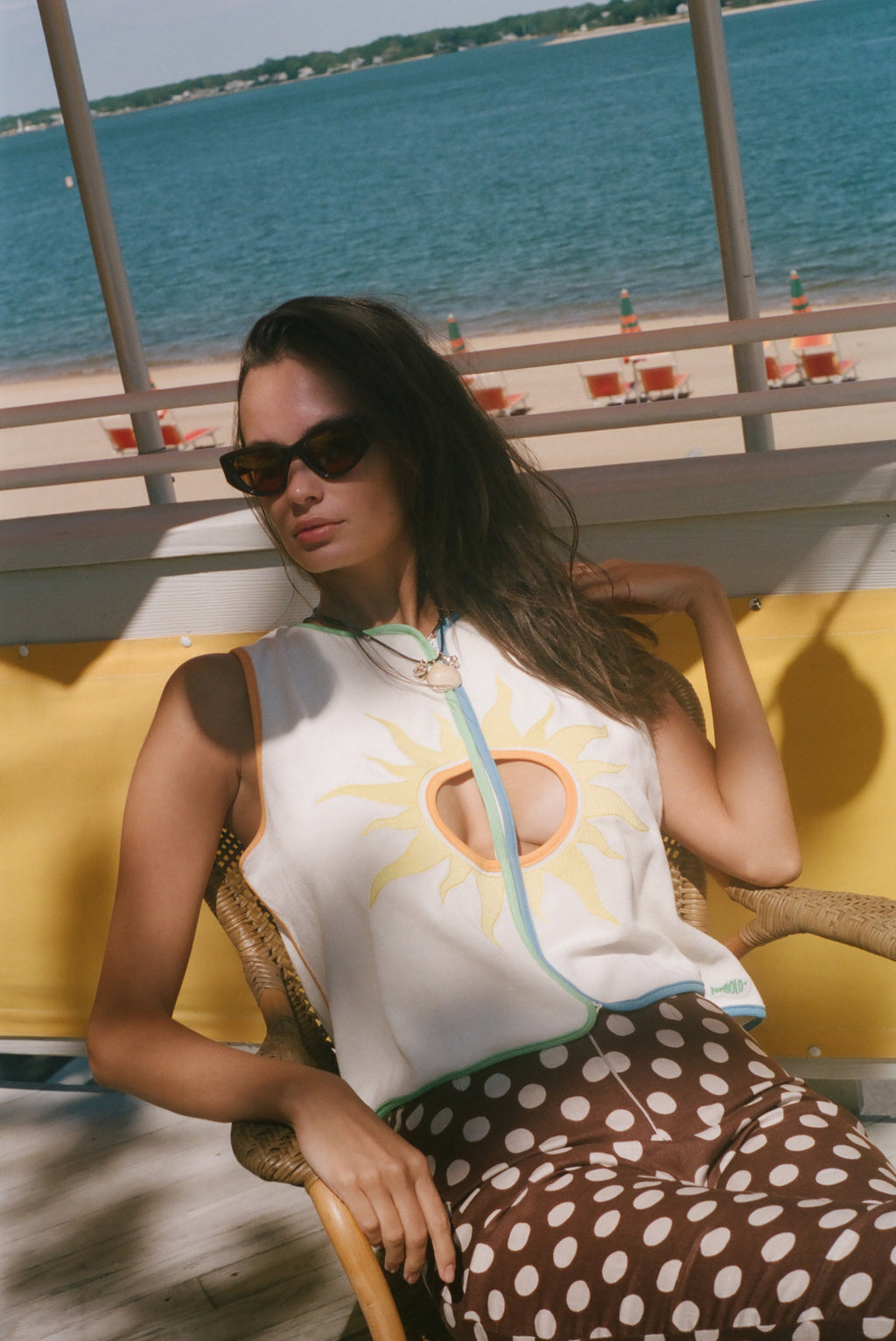 A woman wearing a white sleeveless top with a yellow sun graphic and brown polka-dot pants, seated in a wicker chair with a beach and ocean in the background.