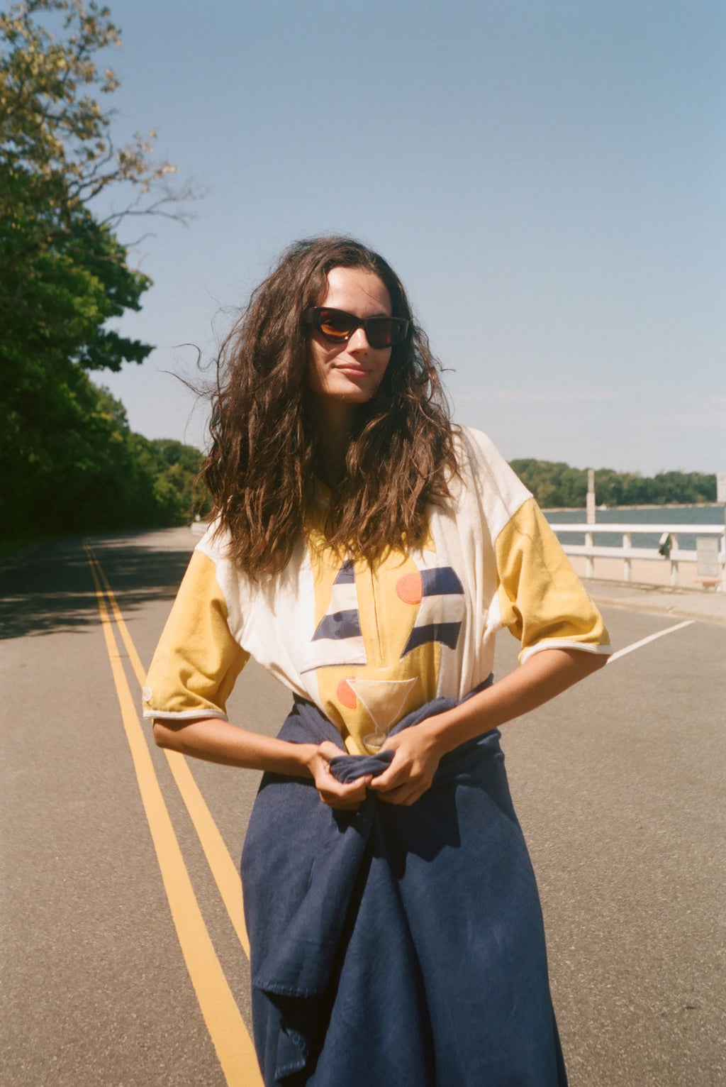 A woman with long, windblown brown hair and dark sunglasses stands on an outdoor road wearing a yellow and white striped shirt.