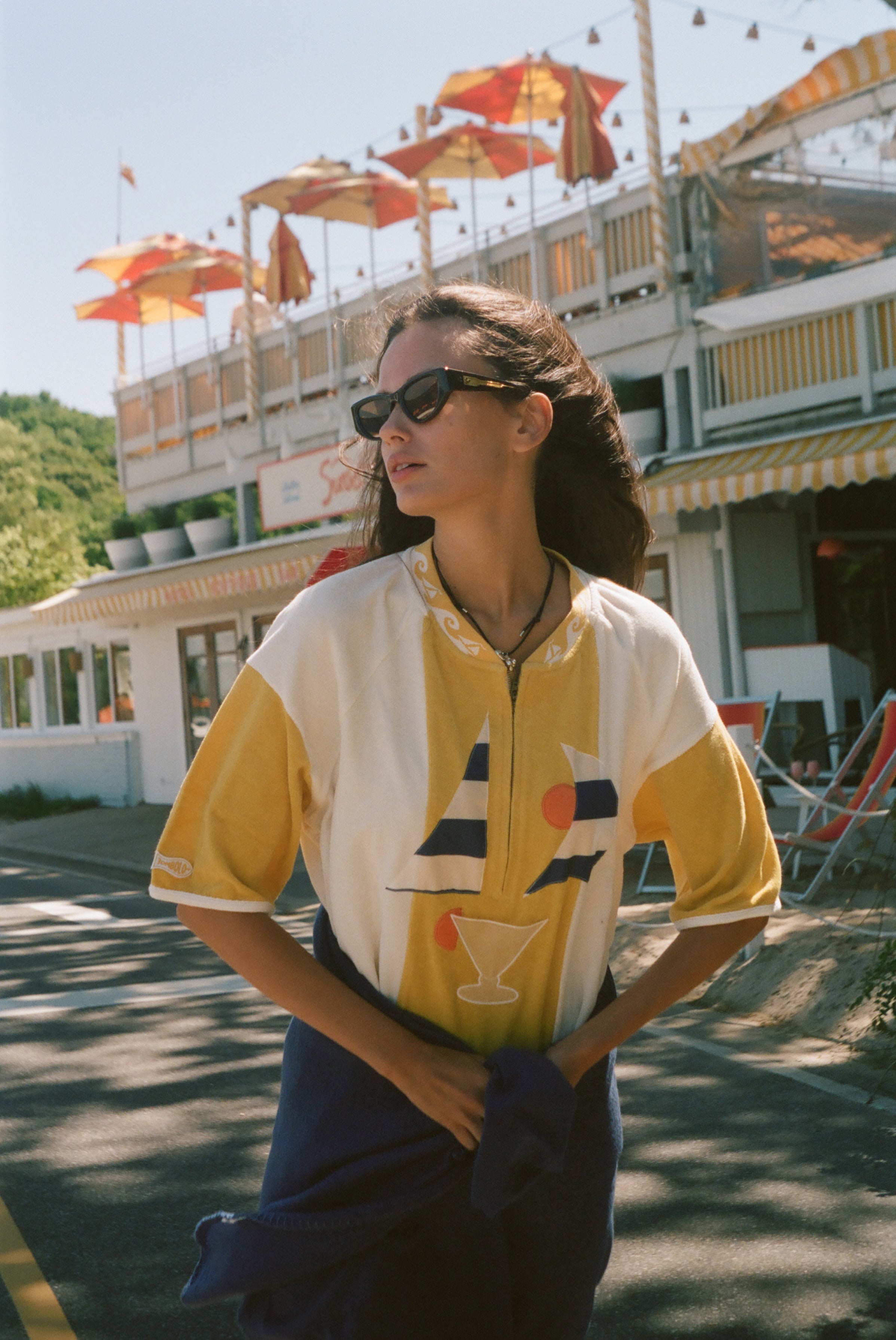 A woman wearing a yellow and white color-blocked short-sleeve shirt with a sailboat and cocktail graphic, styled with dark sunglasses and a navy blue jacket tied around her waist, standing outdoors in front of a building with orange umbrellas.