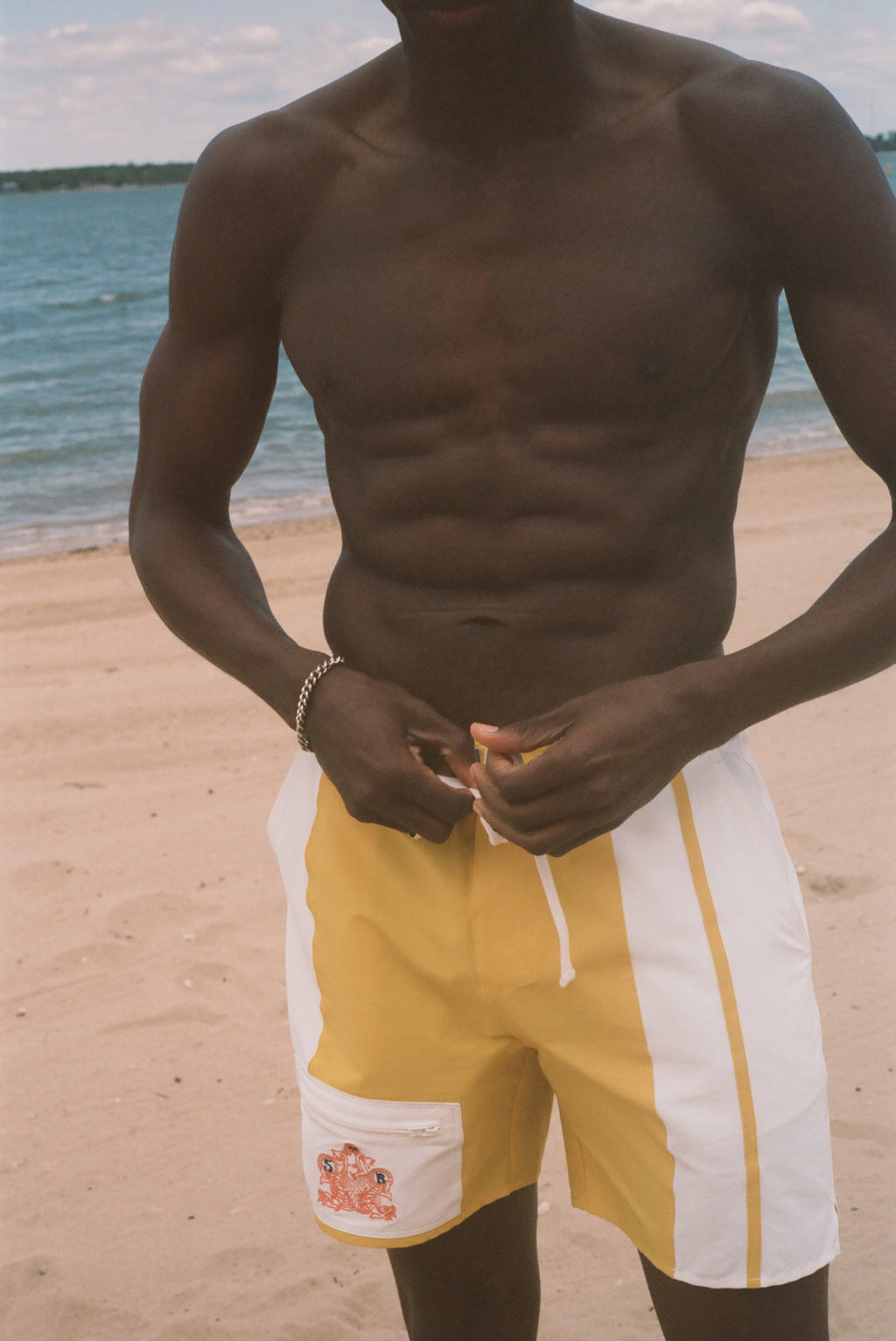 A shirtless man standing on a sandy beach wearing yellow and white swim trunks, with the ocean and sky in the background.