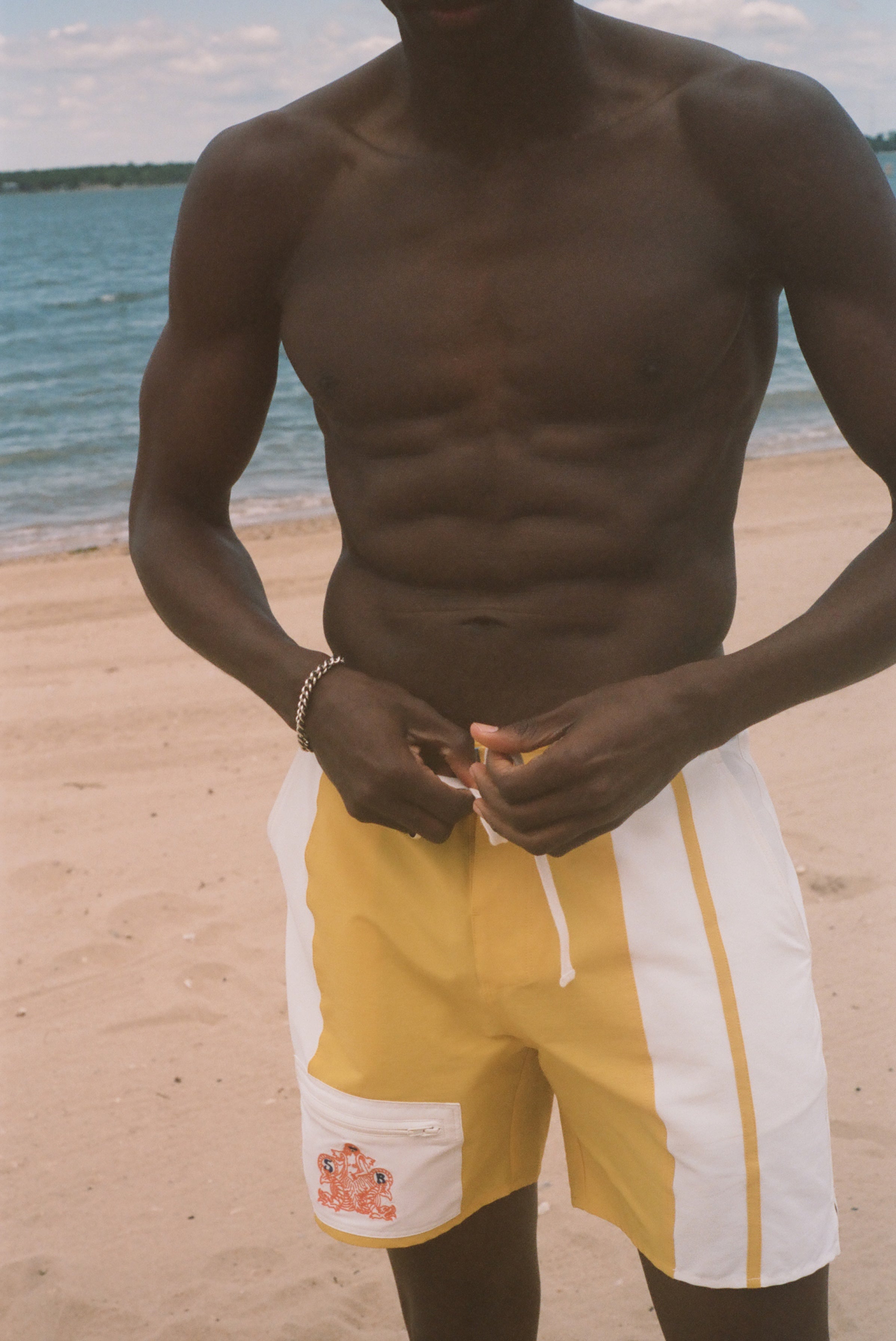 A shirtless man standing on a sandy beach wearing yellow and white swim trunks, with the ocean and sky in the background.