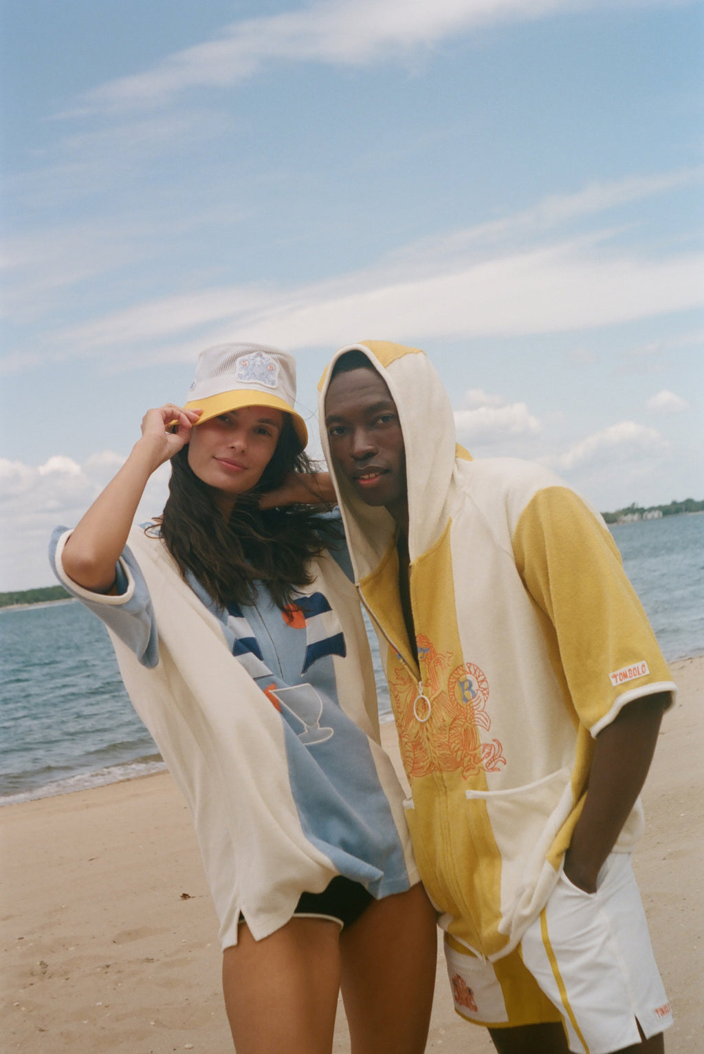 Two models standing on a sandy beach wearing matching yellow and off-white hooded beach poncho-style tops under a blue sky.