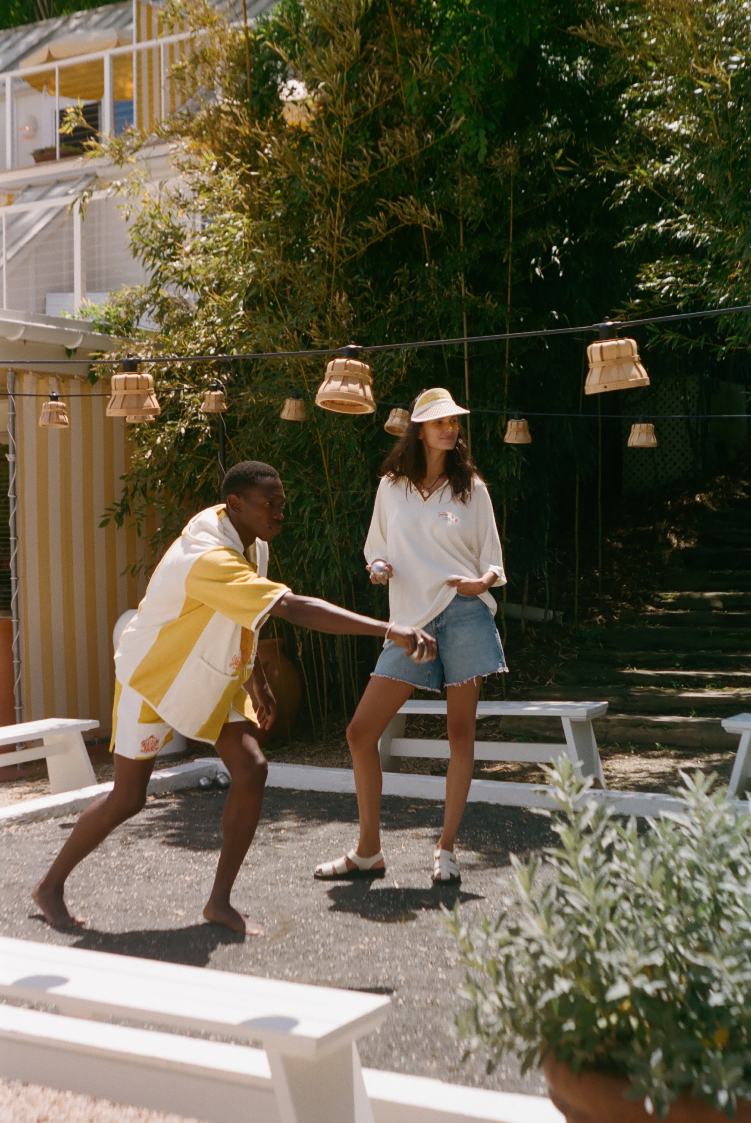 A man and a woman outdoors in a sunny garden or patio setting. The man wears a white and yellow striped shirt and shorts, while the woman wears a white long-sleeve top, denim shorts, and a straw hat.