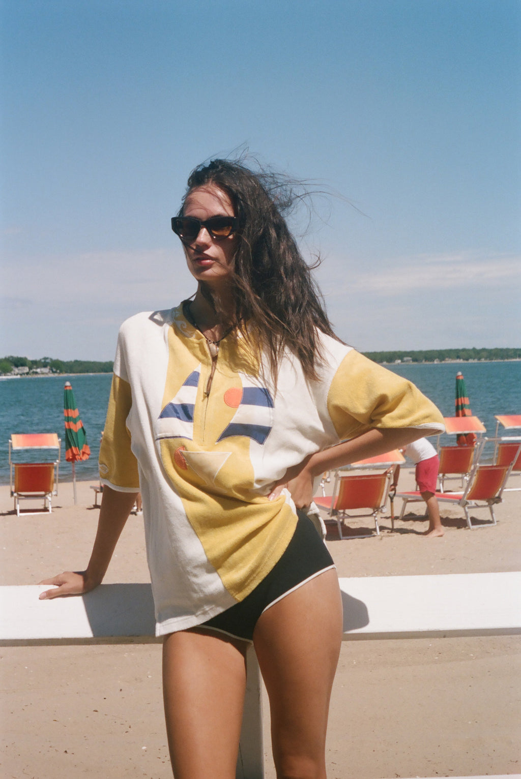A woman stands on a beach wearing an oversized yellow and white graphic t-shirt, black swim trunks, and sunglasses, with the ocean and lounge chairs in the background.