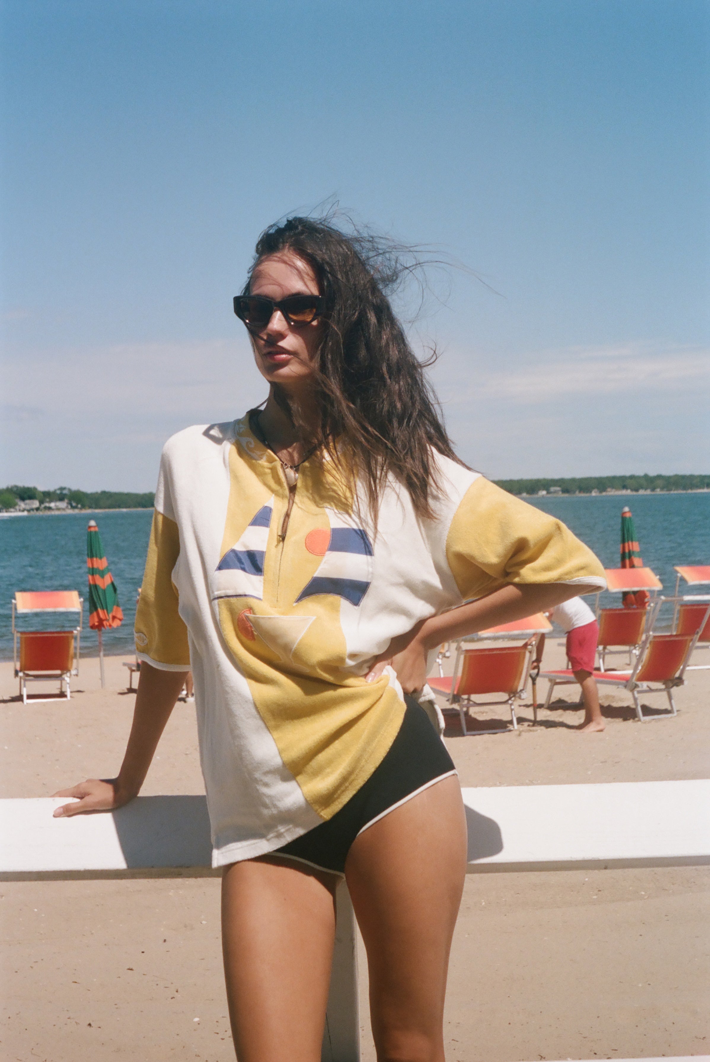 A woman stands on a beach wearing an oversized yellow and white graphic t-shirt, black swim trunks, and sunglasses, with the ocean and lounge chairs in the background.