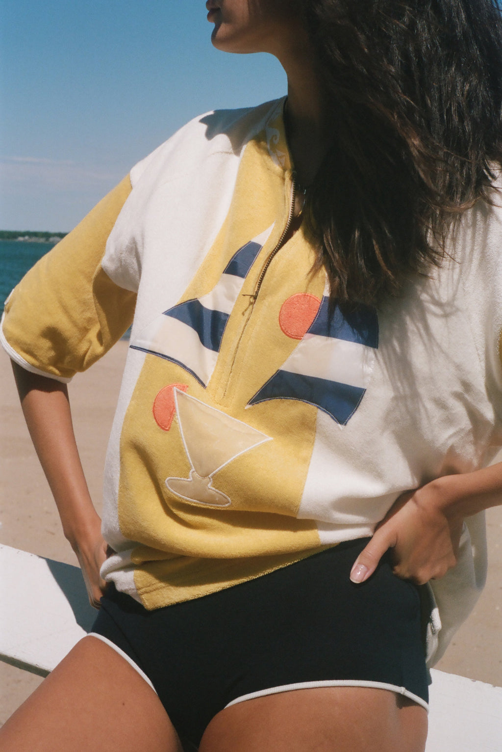 A woman at the beach wearing a yellow and white short-sleeve zip-up shirt with a graphic print of a cocktail glass and blue geometric shapes, paired with black swim shorts.