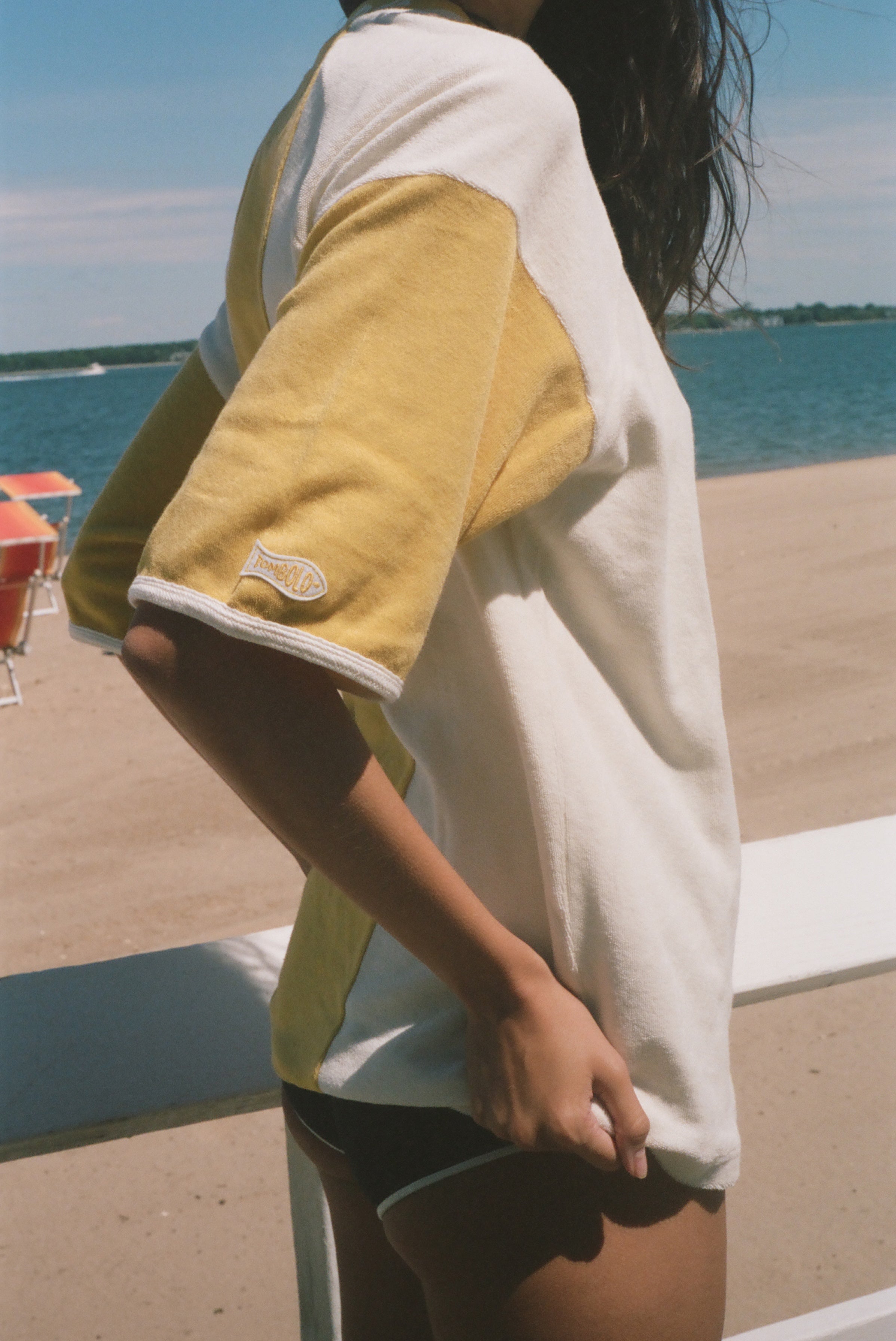 A woman stands on a beach wearing an oversized yellow and white graphic t-shirt, black swim trunks, and sunglasses, with the ocean and lounge chairs in the background.