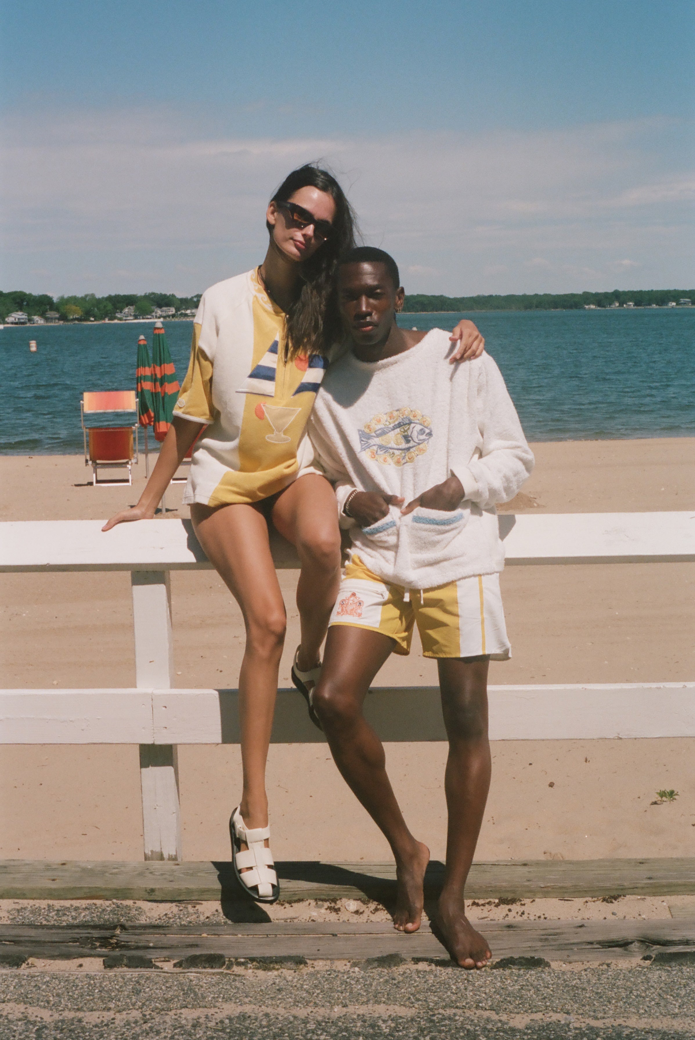 A couple posing in matching yellow and white swimwear and loungewear sets on a beach boardwalk with a white fence, ocean, and sky in the background.