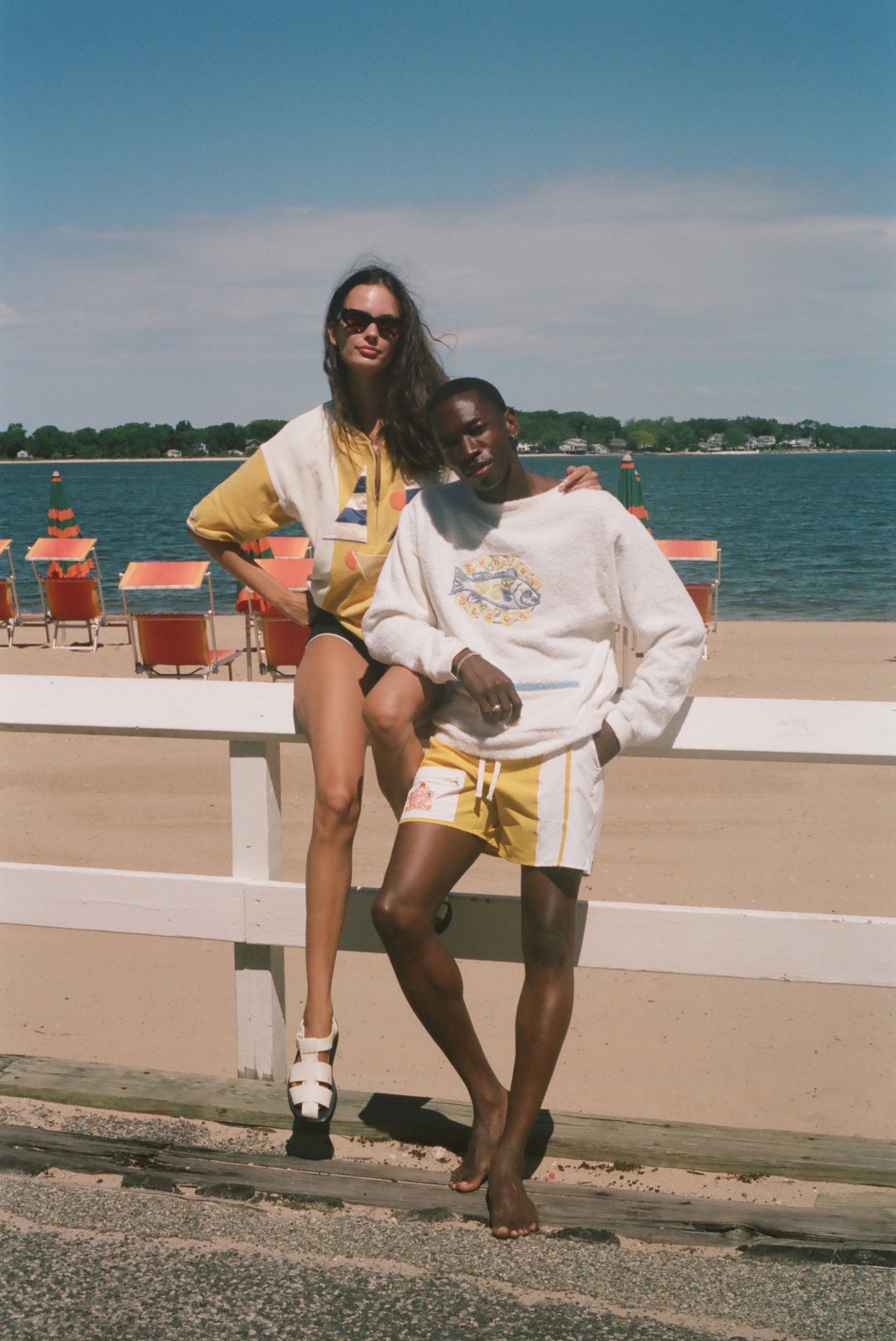 Two models posing on a beach boardwalk in front of a white railing, with the ocean, sand, and beach chairs in the background. The woman wears a yellow and white striped poncho.
