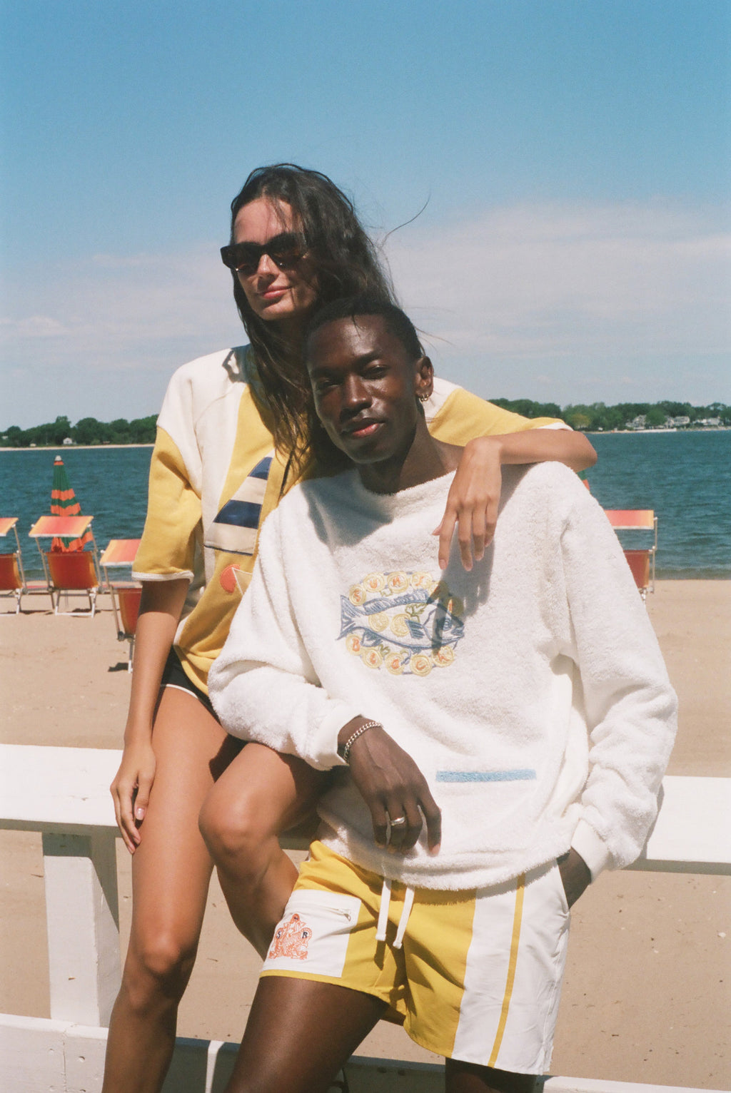 Two models posing on a white beach chair at the beach, wearing yellow and white swimwear and a white sweatshirt with a graphic print.