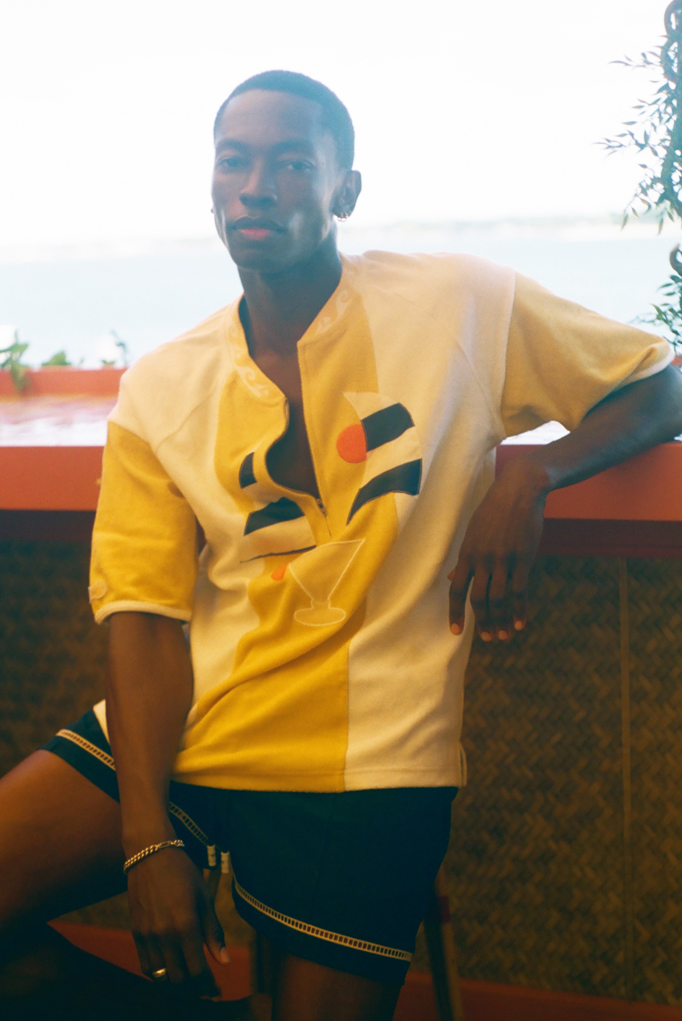 A man wearing a yellow and white short-sleeve button-up shirt with a geometric graphic on the front, paired with dark shorts, leaning against a wall in an outdoor setting.
