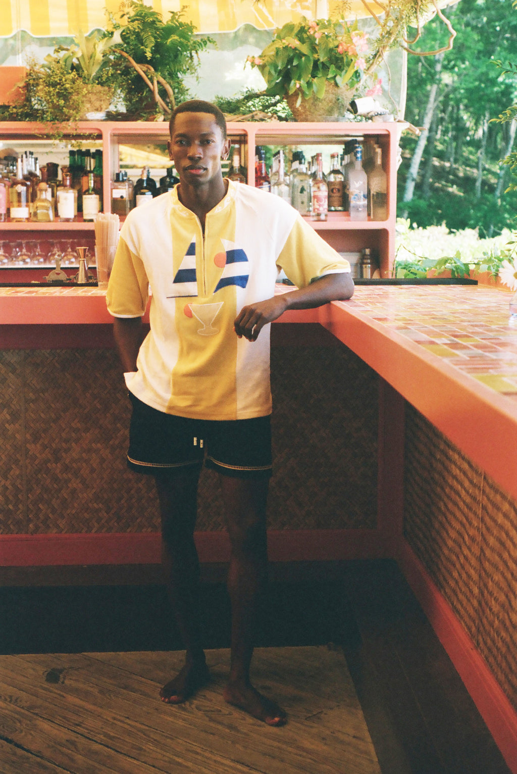 A man wearing a yellow and white short-sleeve athletic jersey with blue and red accents, paired with black athletic shorts, standing in a tropical bar environment.