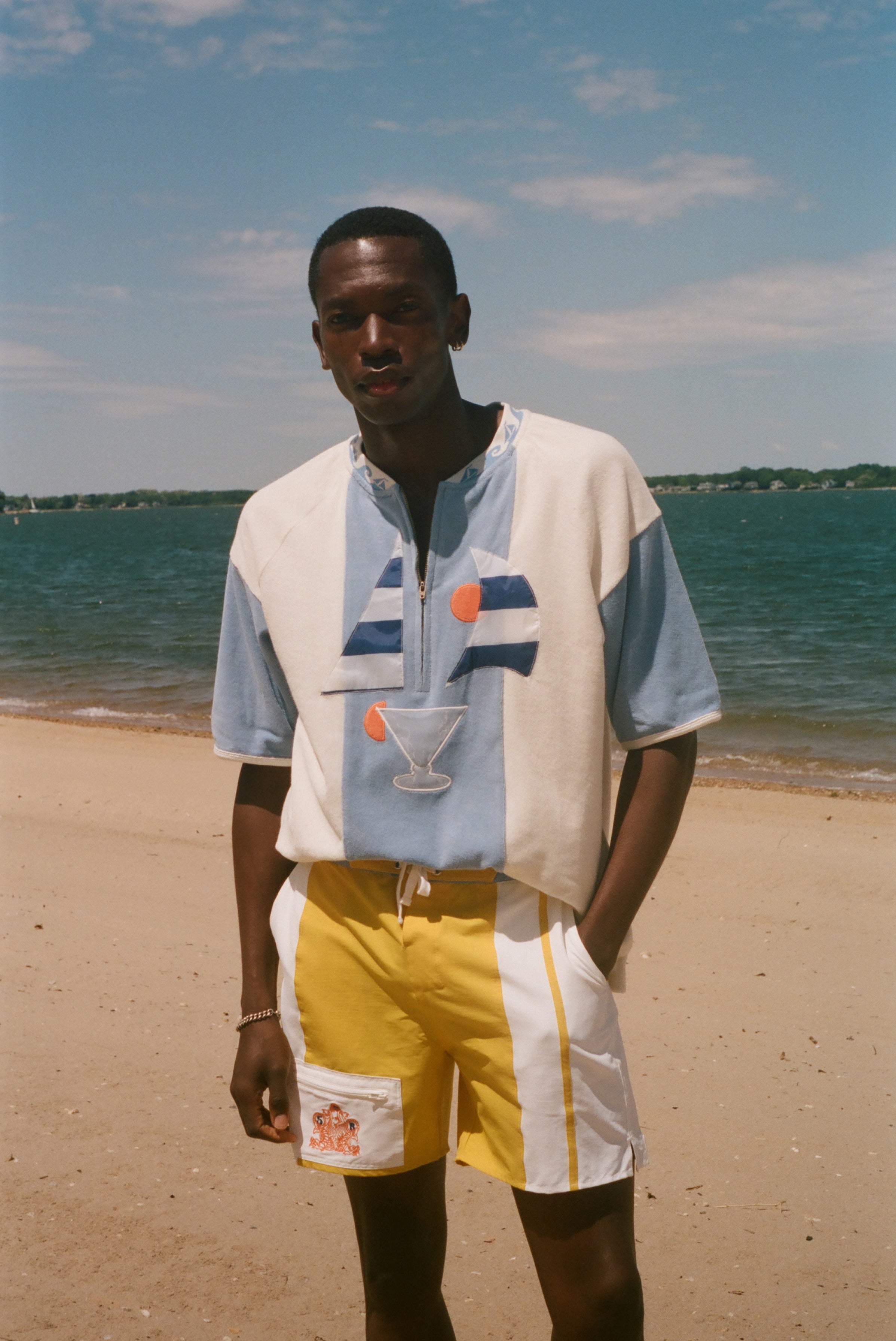 A man standing on a sandy beach wearing a color-blocked short-sleeve shirt with a light blue and white base, a blue and orange sailboat graphic, and a cocktail glass print, paired with yellow and white shorts.