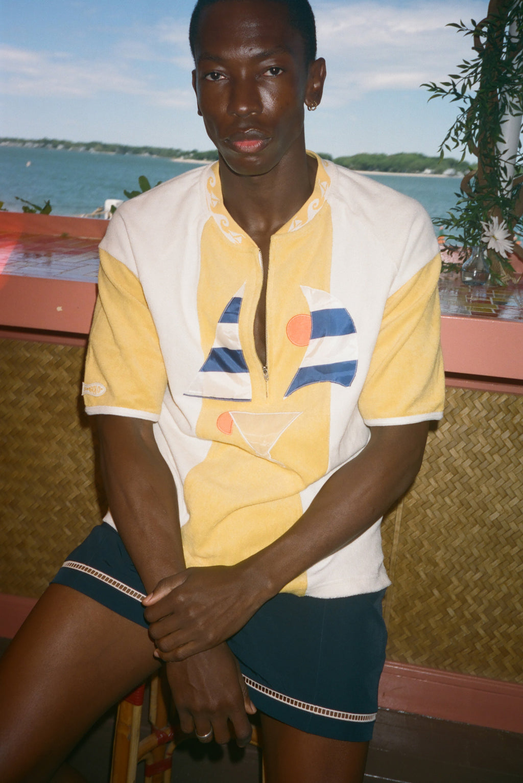 A man wearing a yellow and white color-blocked short-sleeve polo shirt with a geometric print, paired with dark shorts, seated in front of a scenic ocean background.