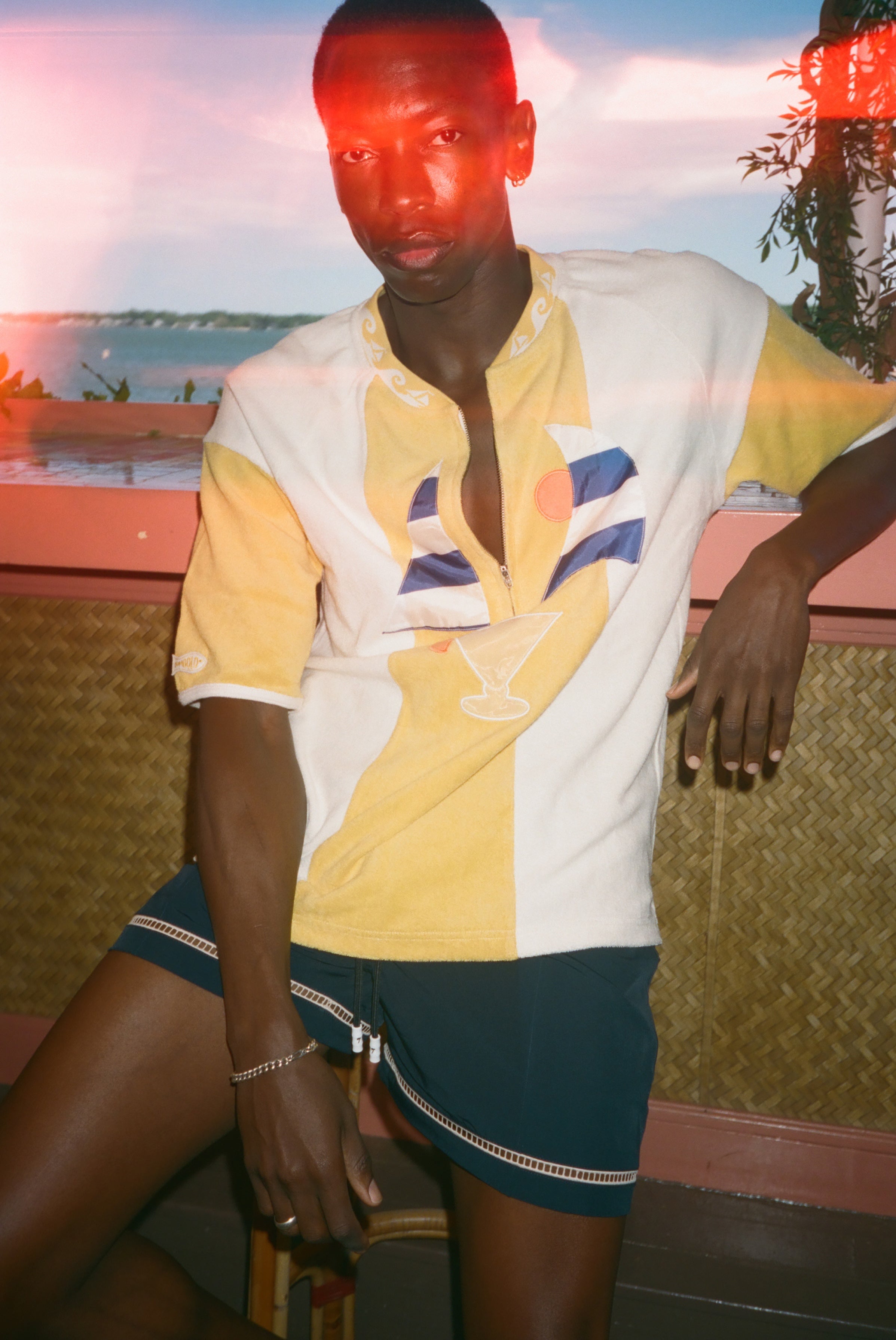 A man wearing a yellow and white color-blocked polo shirt with a cocktail graphic on the front, paired with dark teal shorts, posed against a tropical sunset background.