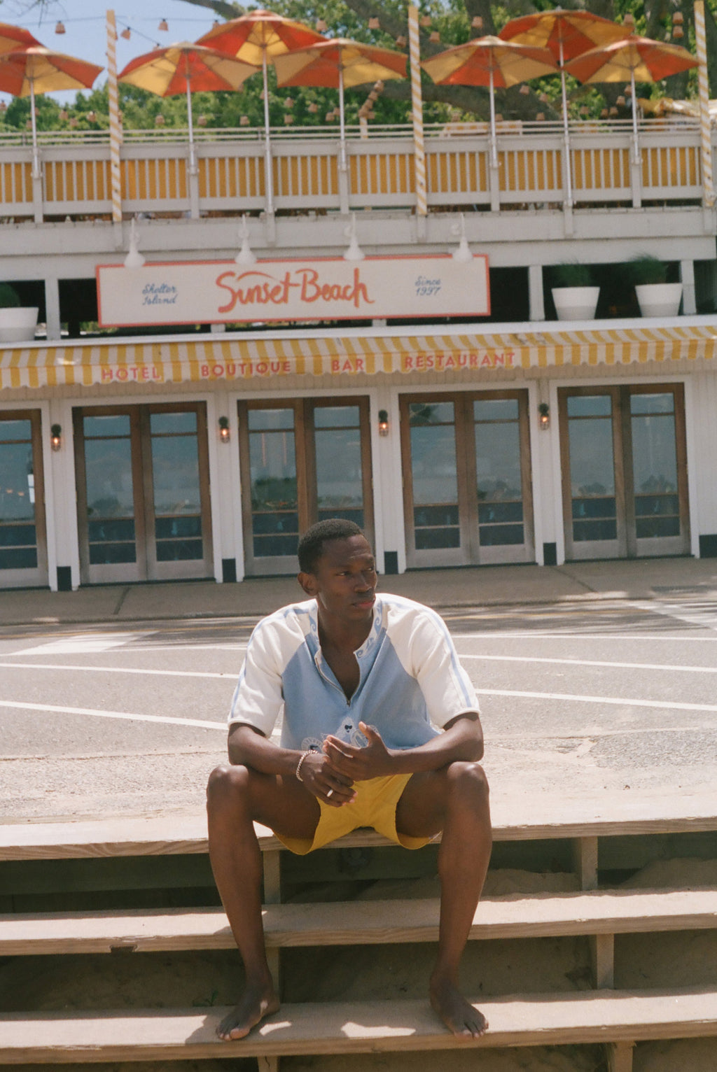 A man sits barefoot on concrete steps in front of a building labeled 'Sunset Beach' and 'Hotel, Restaurant & Bar'. He is wearing a white and light blue short-sleeve shirt with yellow shorts.