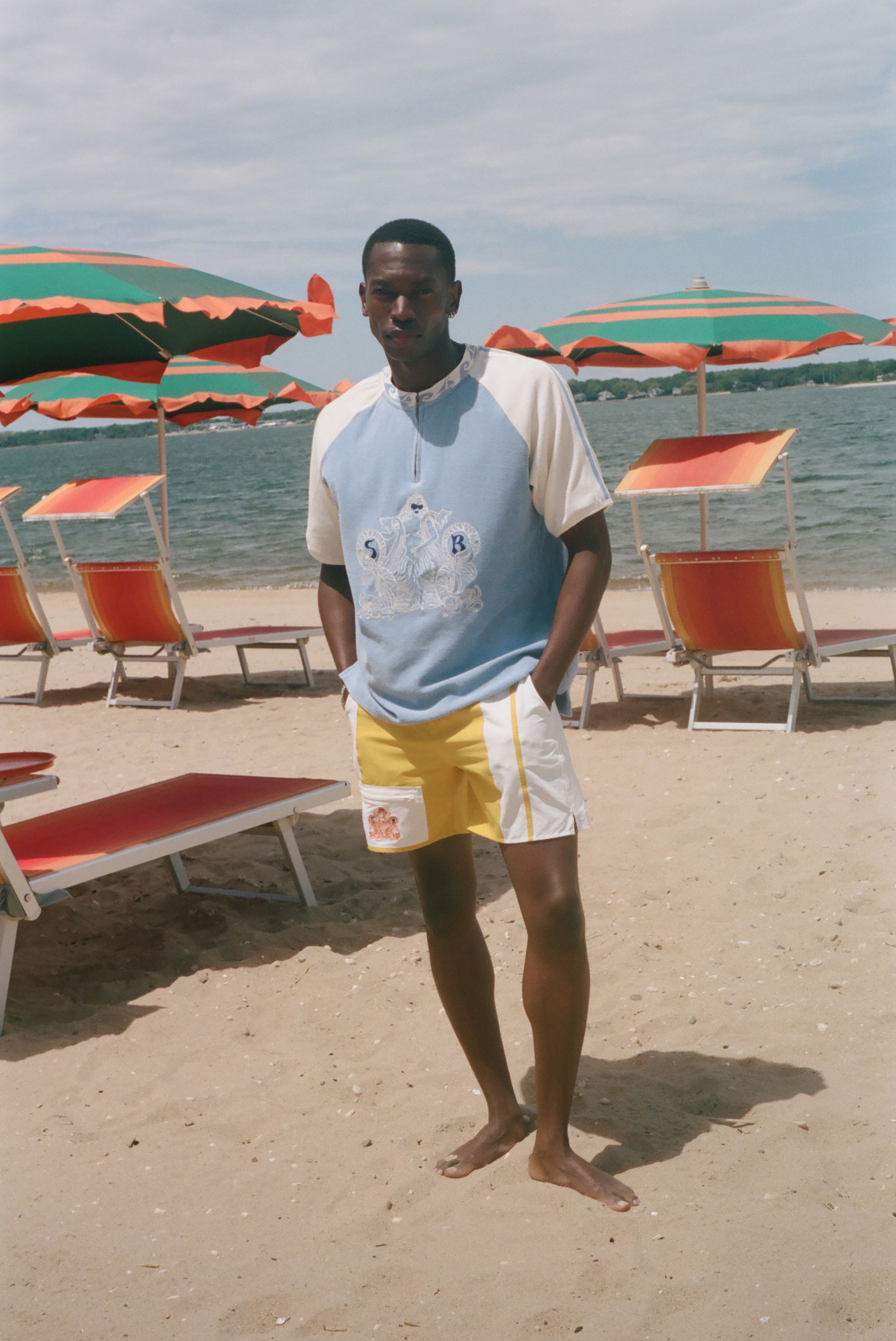 A man stands on a sandy beach wearing a light blue and white raglan t-shirt with a graphic print, yellow and white swim trunks, and standing barefoot in front of beach umbrellas and lounge chairs.